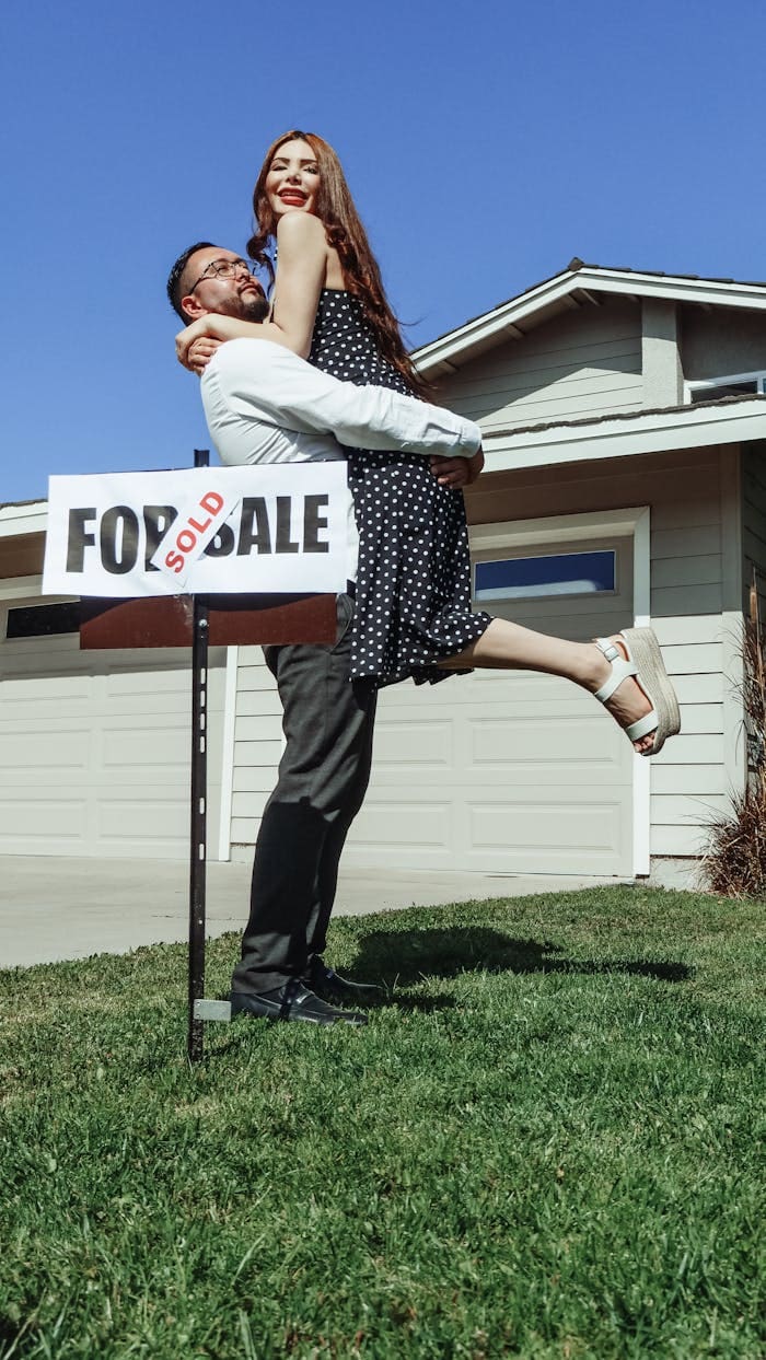A joyful couple celebrates buying their new house with a Sold sign.
